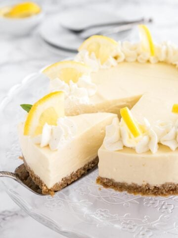 A lemon cream pie on a glass serving platter topped with whipped cream and lemon wedges. A cake server is lifting out a slice.
