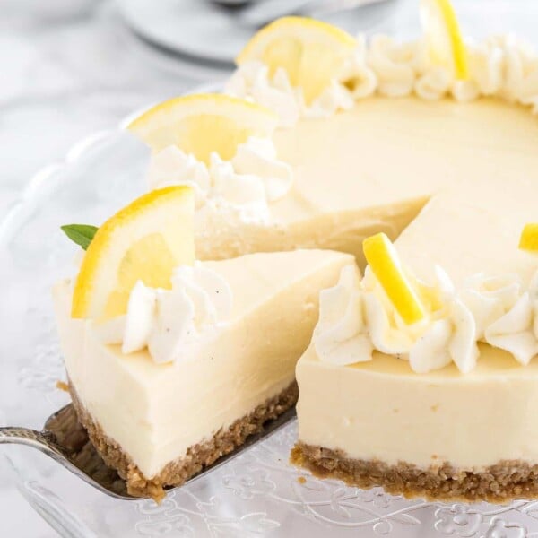 A lemon cream pie on a glass serving platter topped with whipped cream and lemon wedges. A cake server is lifting out a slice.