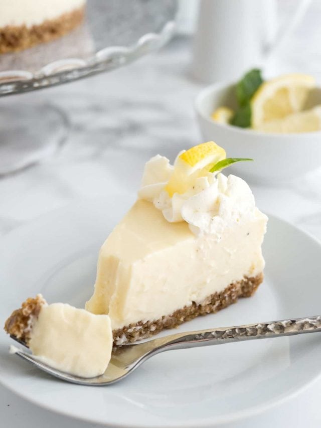 A slice of lemon cream pie on a white plate with a fork on a marble surface. There\'s a glass platter with the rest of the cake and a small bowl of lemon slices in the background. The fork has taken out a piece of the pie.