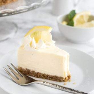 A slice of lemon cream pie on a white plate with a fork on a marble surface. There's a glass platter with the rest of the cake and a small bowl of lemon slices in the background.