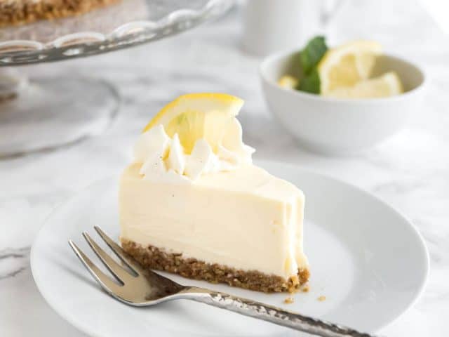 A slice of lemon cream pie on a white plate with a fork on a marble surface. There\'s a glass platter with the rest of the cake and a small bowl of lemon slices in the background.