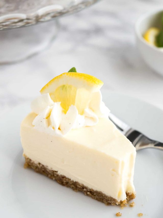 Close-up of A slice of lemon cream pie on a white plate with a fork on a marble surface. There\'s a glass platter and a small bowl of lemon slices in the background.