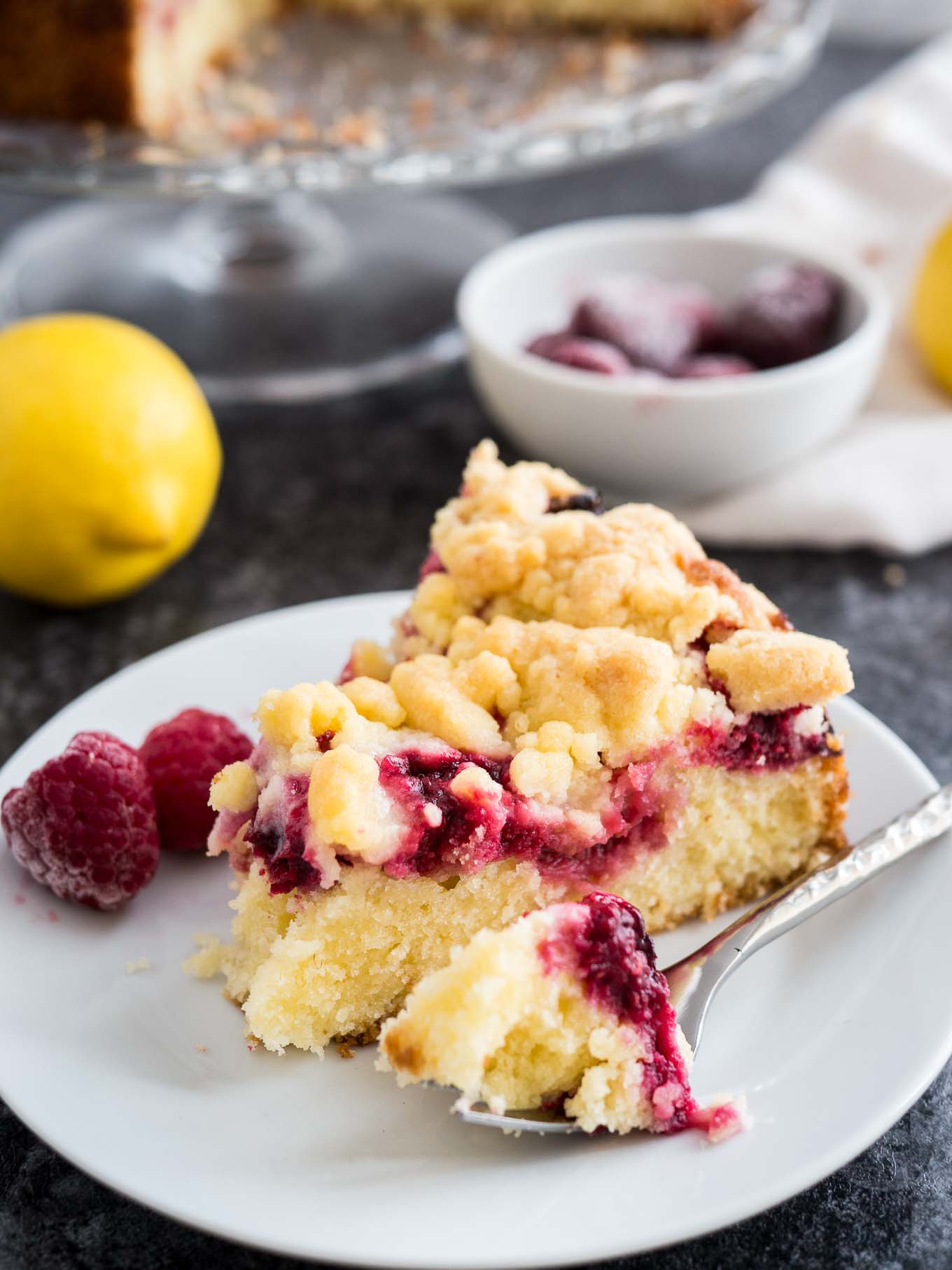 A slice of lemon raspberry cake topped with streusel on a white pate with a fork garnished with raspberries. There is a lemon, a white bowl of frozen raspberries and a glass serving platter with the rest of the cake in the background.