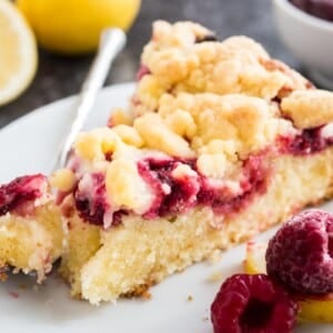 A slice of lemon raspberry cake with streusel topping on a white plate with a fork with a small piece of the cake on it, garnished with lemon and raspberries.