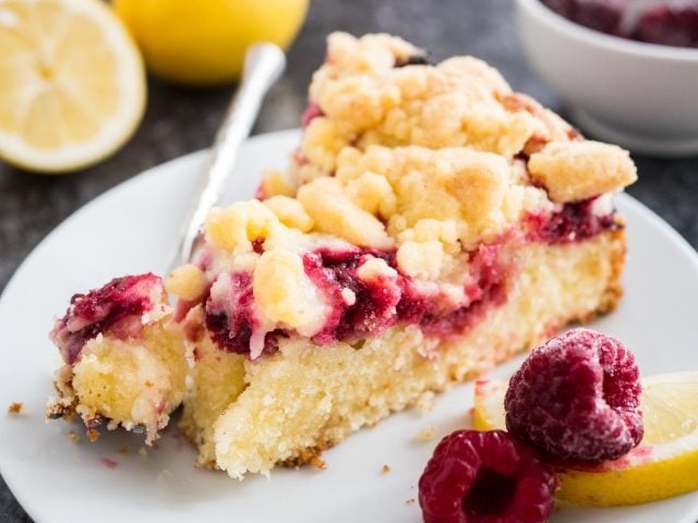 A slice of lemon raspberry cake with streusel topping on a white plate with a fork with a small piece of the cake on it, garnished with lemon and raspberries.