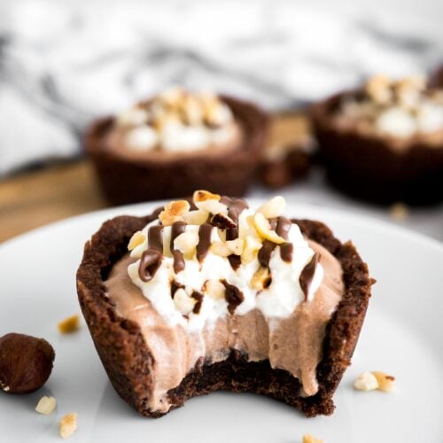 A white plate with a nutella mousse cookie cup with a bite taken out of it garnished with hazelnuts. There are more cookie cups in the background.