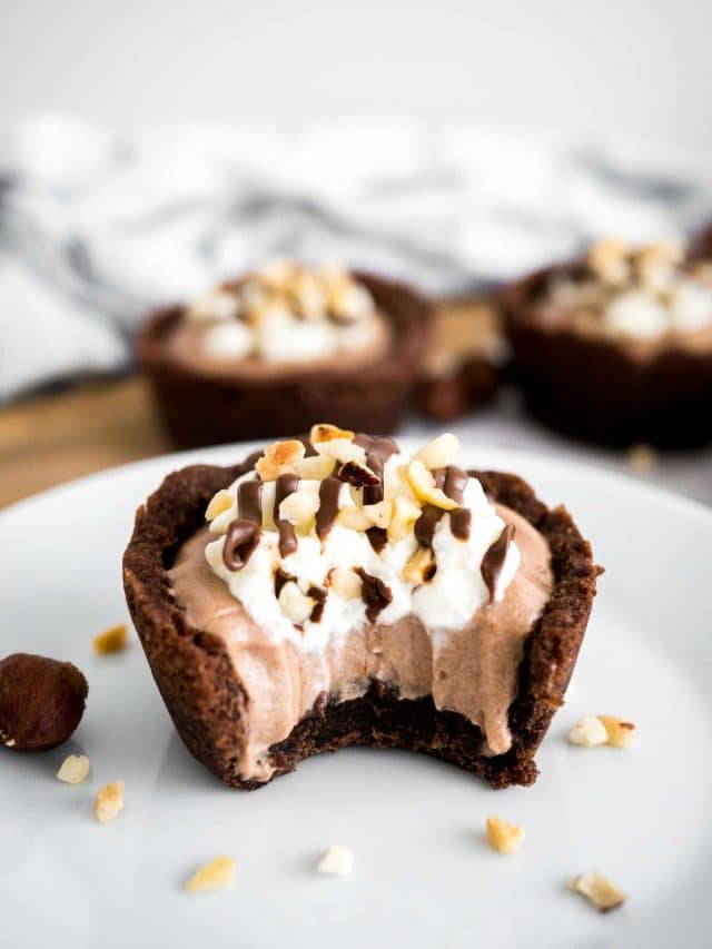 A white plate with a nutella mousse cookie cup with a bite taken out of it garnished with hazelnuts. There are more cookie cups in the background.