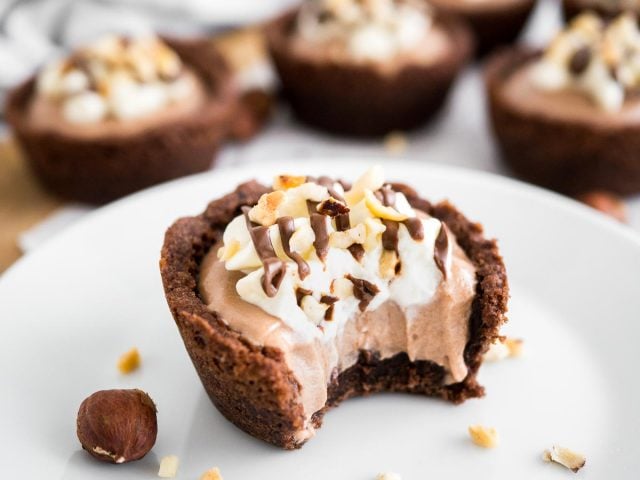 A white plate with a nutella mousse cookie cup with a bite taken out of it garnished with hazelnuts. There are more cookie cups in the background.