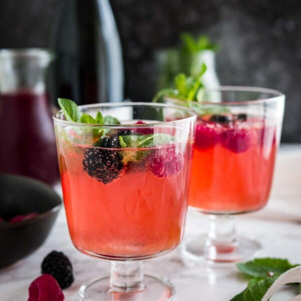 Two glasses of sparkling berry champagne cocktail with raspberries and mint on a marble surface garnished with raspberries and mint. There's a black bowl and a carafe of dark red juice in the background.