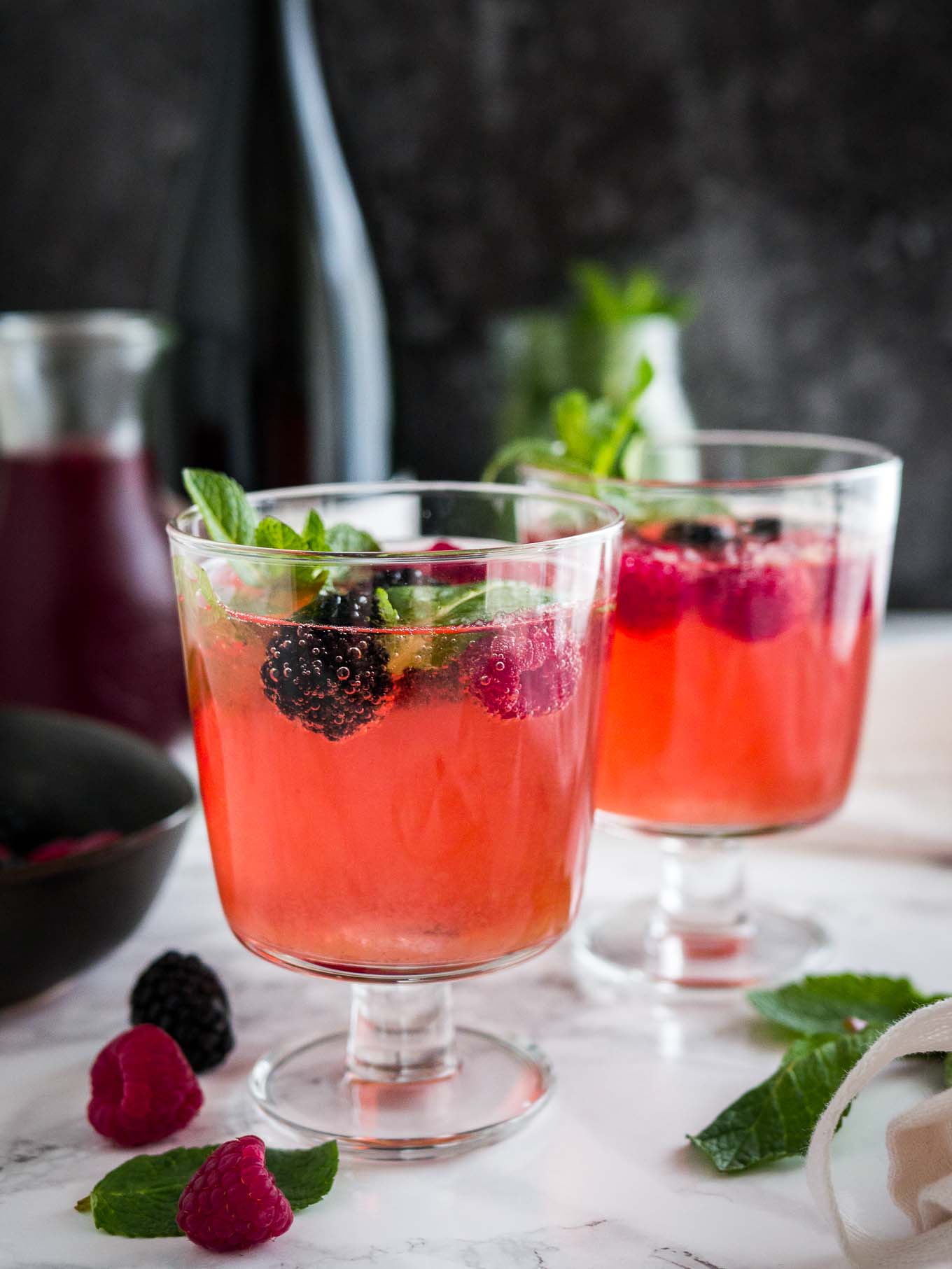 Two glasses of sparkling berry champagne cocktail with raspberries and mint on a marble surface garnished with raspberries and mint. There\'s a black bowl and a carafe of dark red juice in the background.