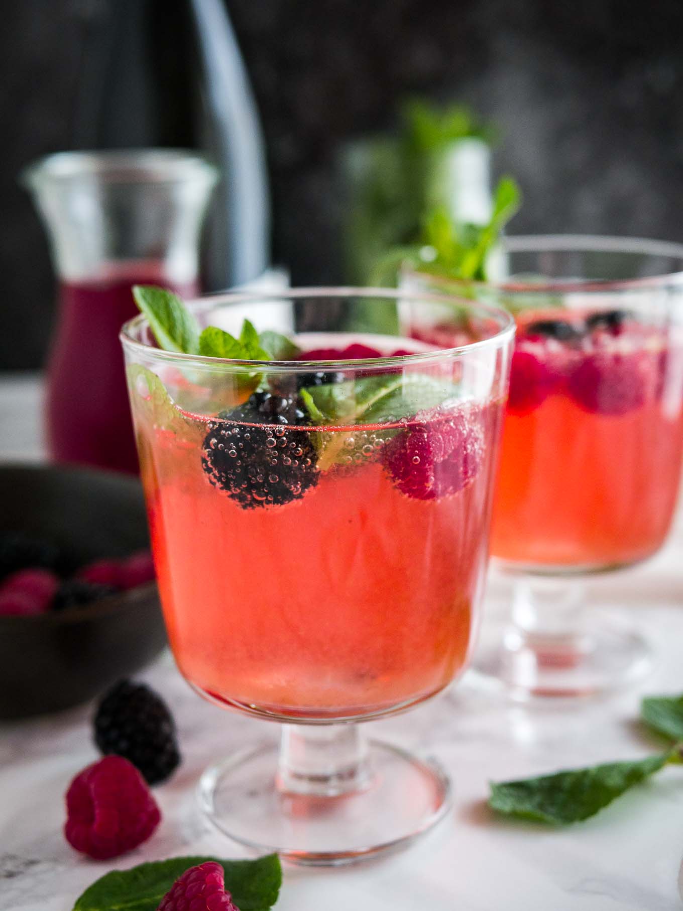 Two glasses of sparkling berry champagne cocktail with raspberries and mint on a marble surface garnished with raspberries and mint. There\'s a black bowl and a carafe of dark red juice in the background.