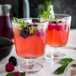 Two glasses of sparkling berry champagne cocktail with raspberries and mint on a marble surface garnished with raspberries and mint. There's a black bowl and a carafe of dark red juice in the background.