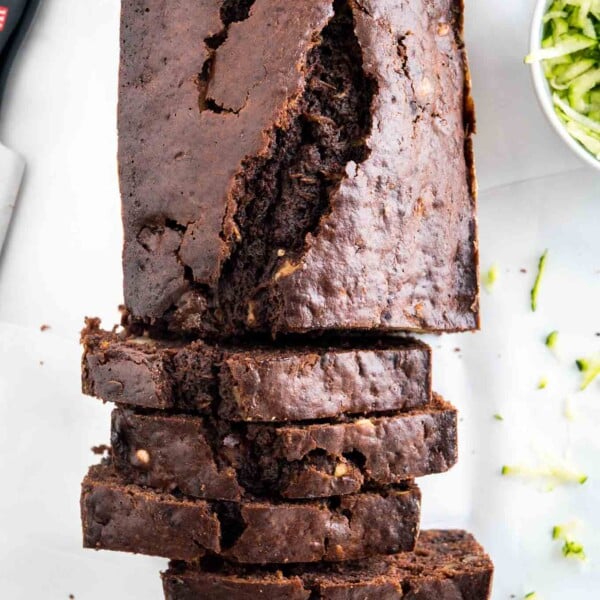 Top-down shot of a chocolate zucchini bread with some slices cut off and standing in front of it. There's a knife and a white bowl of shredded zucchini next to it.