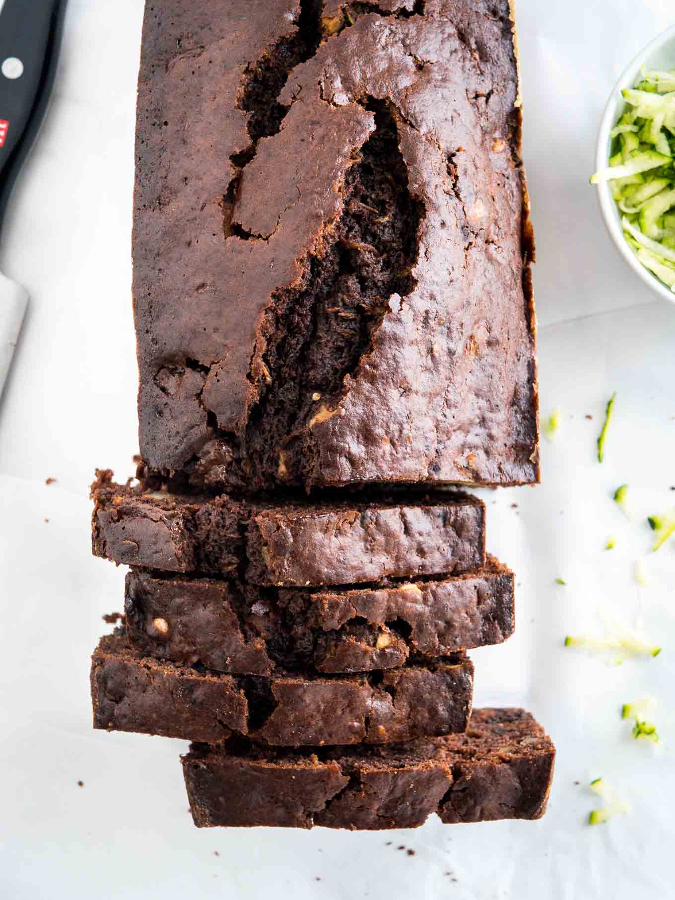 Top-down shot of a chocolate zucchini bread with some slices cut off and standing in front of it. There\'s a knife and a white bowl of shredded zucchini next to it.
