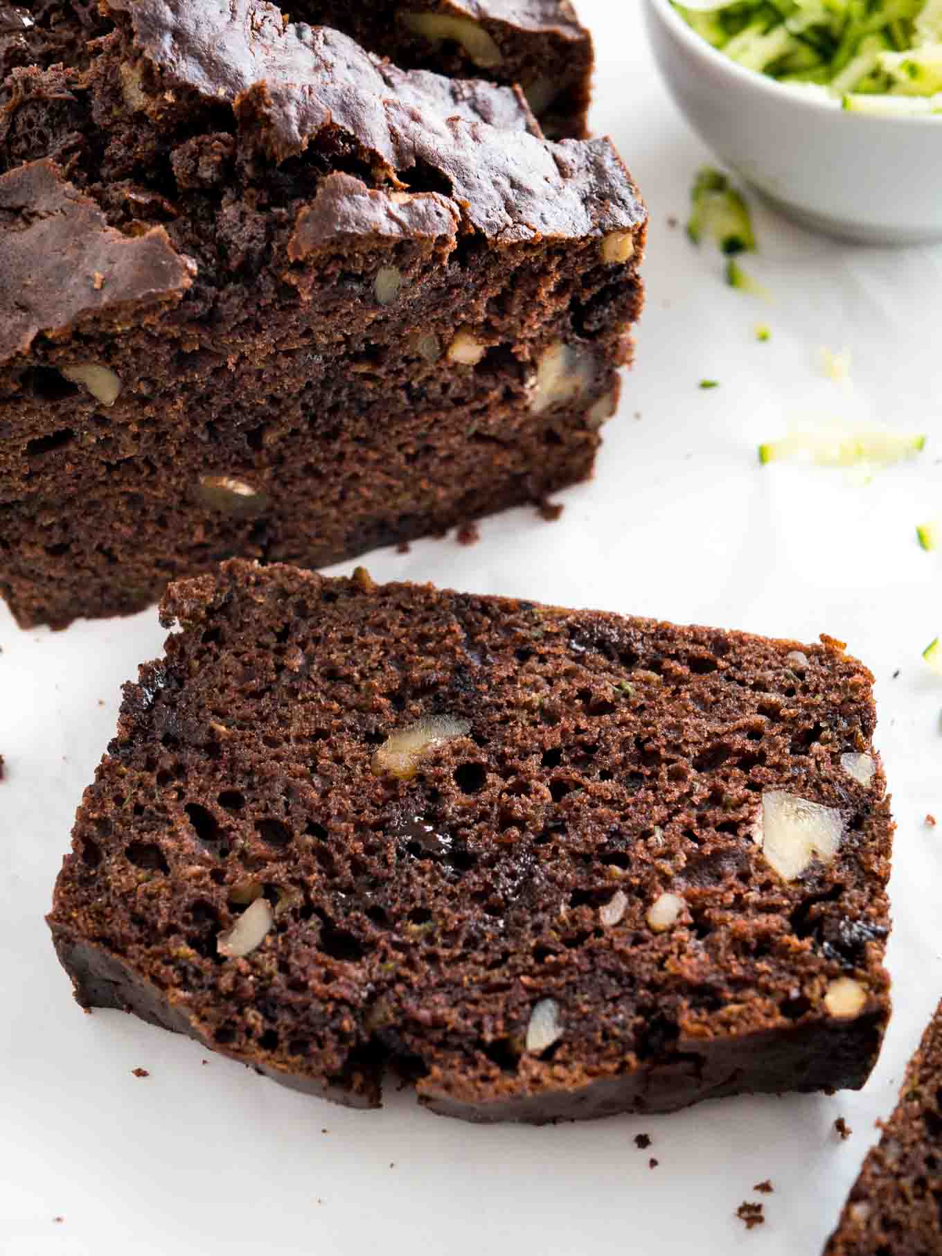 Close-up of a loaf of chocolate zucchini bread with two slices cut off, one standing one lying in front. There\'s a white bowl of shredded zucchini next to it.