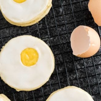 Several easter sugar cookies with lemon curd (resembling a sunny side up egg) on a black cooling rack with some eggshells next to it.