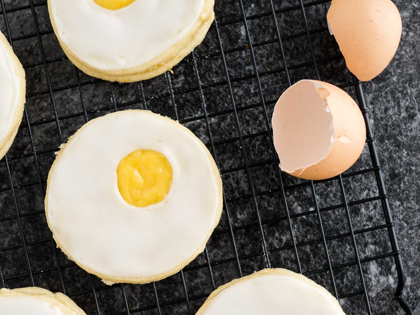 Several easter sugar cookies with lemon curd (resembling a sunny side up egg) on a black cooling rack with some eggshells next to it.