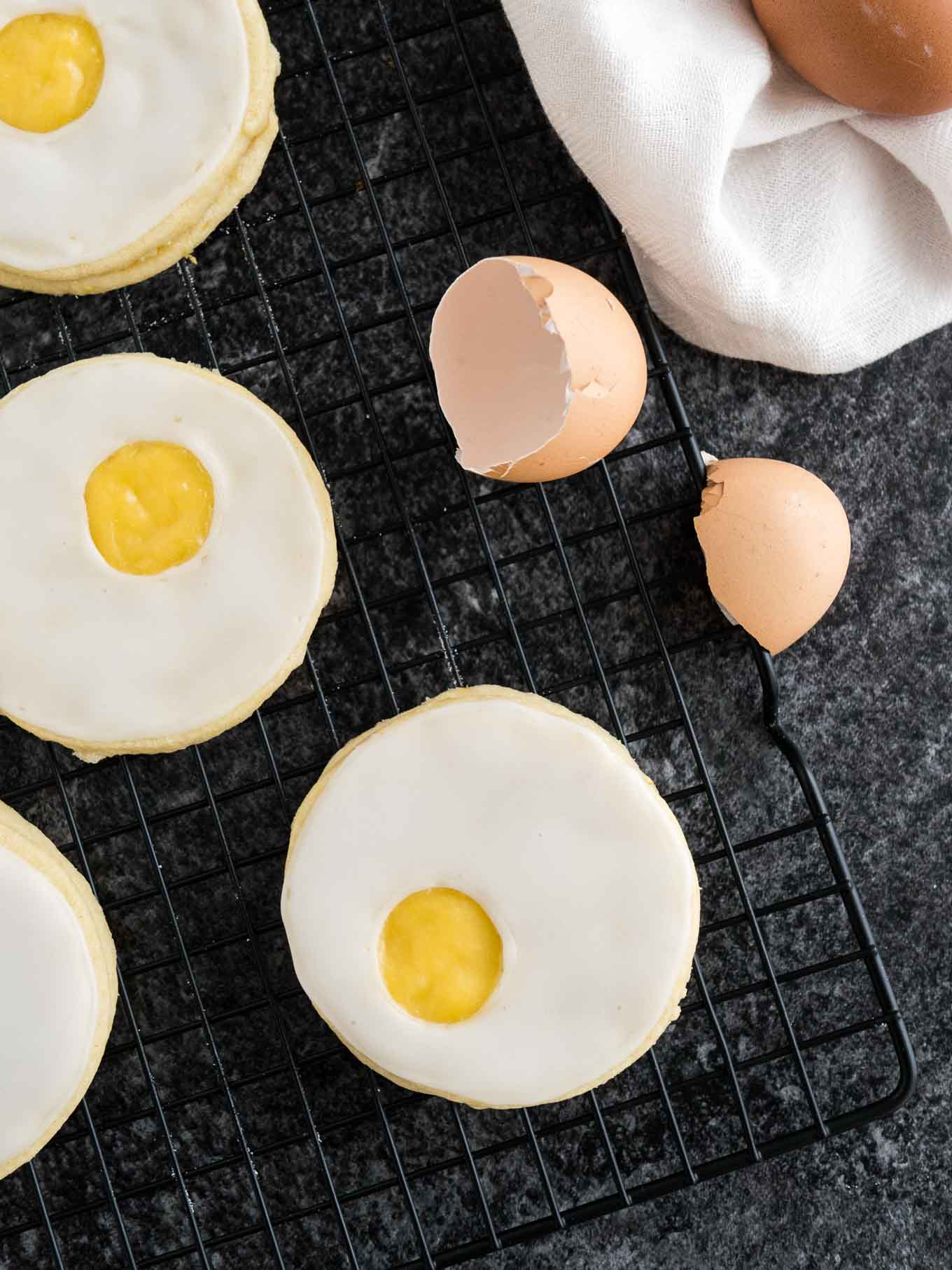 Several easter sugar cookies with lemon curd (resembling a sunny side up egg) on a black cooling rack with some eggshells and a white dishtowel next to it.