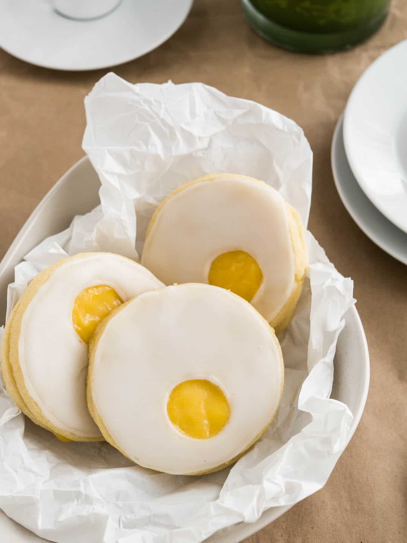 A bowl, lined with parchment paper, with 3 easter sugar cookies with lemon curd (resembling a sunny side up egg) on brown parchment paper next to a stack of plates.