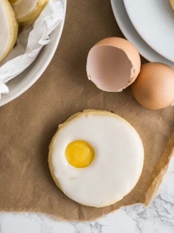 An easter sugar cookie (resembling a sunny side up egg) on parchment paper next to eggshells, a stack of plates and a bowl lined with parchment paper with more of the cookies.