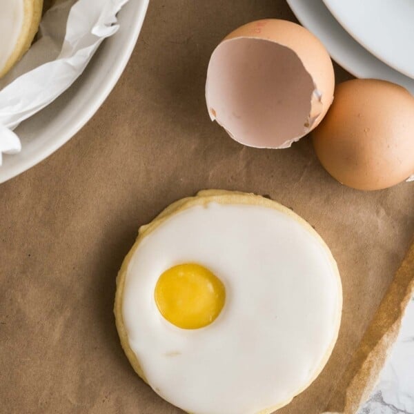 An easter sugar cookie (resembling a sunny side up egg) on parchment paper next to eggshells, a stack of plates and a bowl lined with parchment paper with more of the cookies.