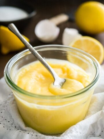 Close-up of a glass jar of lemon curd with a spoon in it on a white dishtowel. There are halved lemons and eggshells in the background.