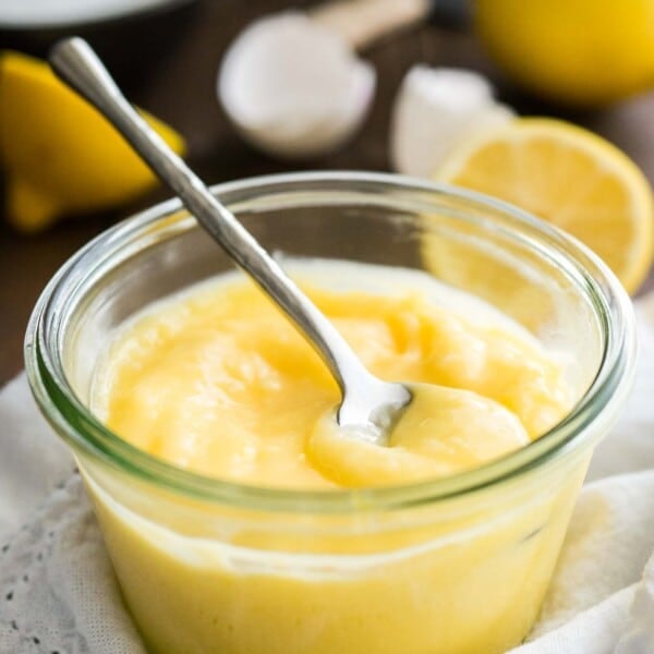 Close-up of a glass jar of lemon curd with a spoon in it on a white dishtowel. There are halved lemons and eggshells in the background.