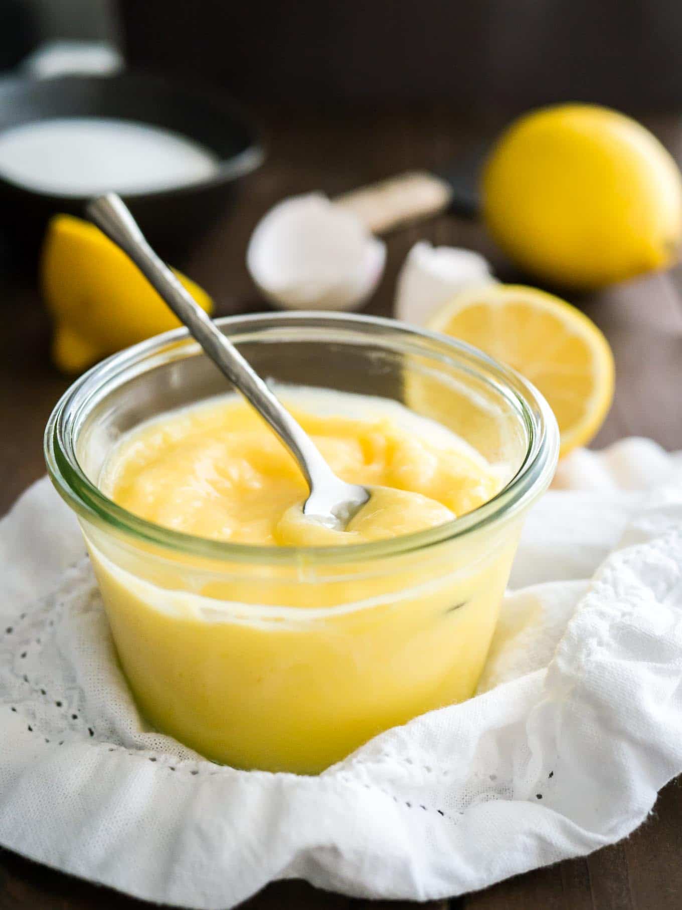 A glass jar of lemon curd with a spoon in it on a white dishtowel. There are halved lemons and eggshells in the background.