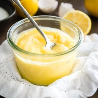 A glass jar of lemon curd with a spoon in it on a white dishtowel. There are halved lemons and eggshells in the background.