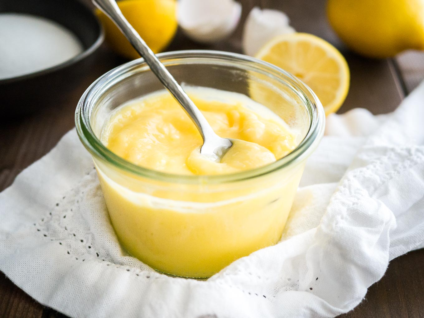 A glass jar of lemon curd with a spoon in it on a white dishtowel. There are halved lemons and eggshells in the background.