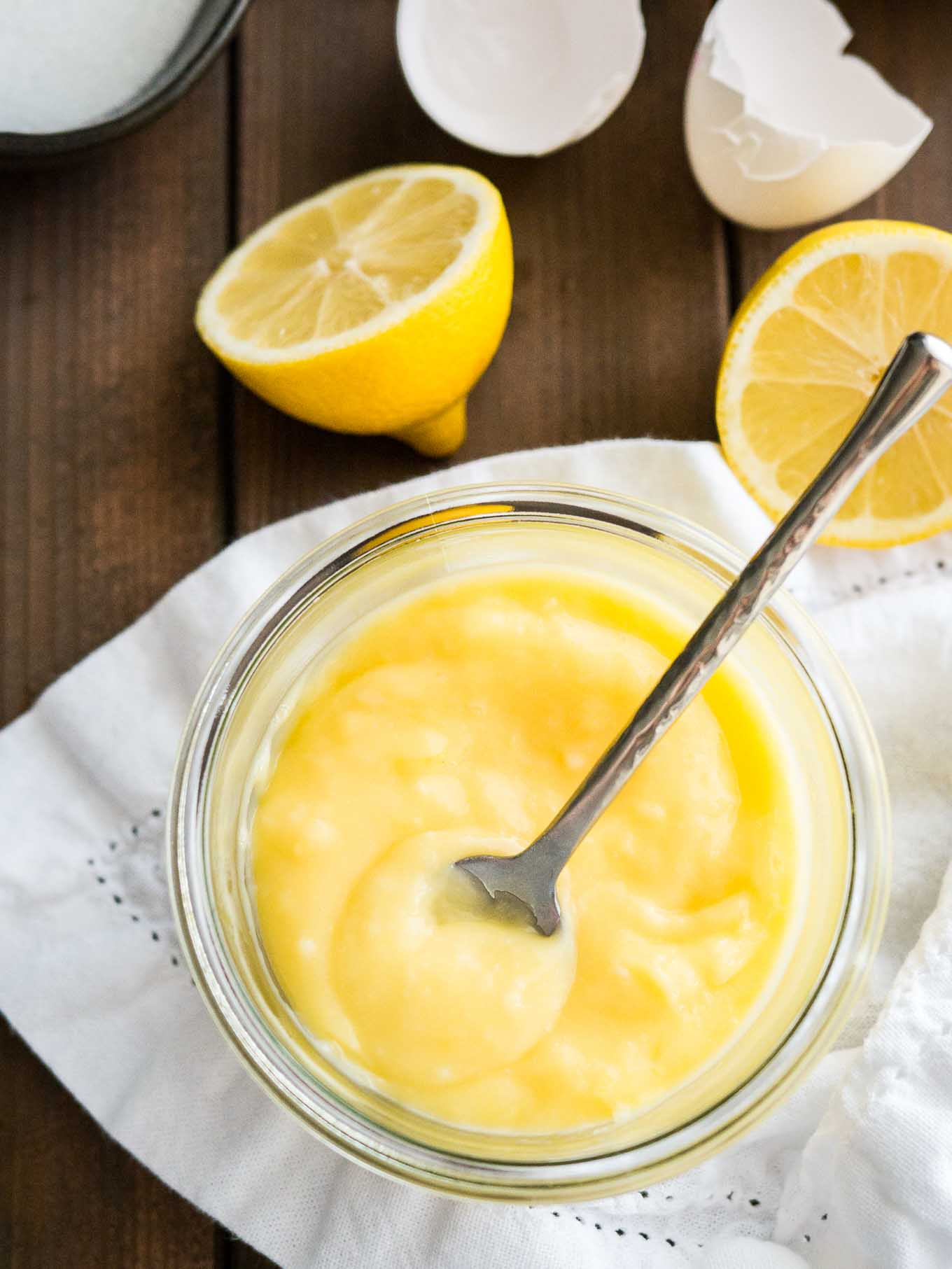 Top-down shot of a glass jar of lemon curd with a spoon in it on a white dishtowel. There are halved lemons and eggshells next to it.