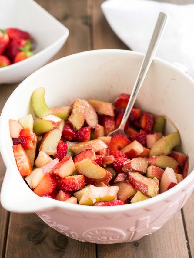 cut strawberries and rhubarb in a pink bowl with a fork in it, fresh strawberries in a bowl in the background