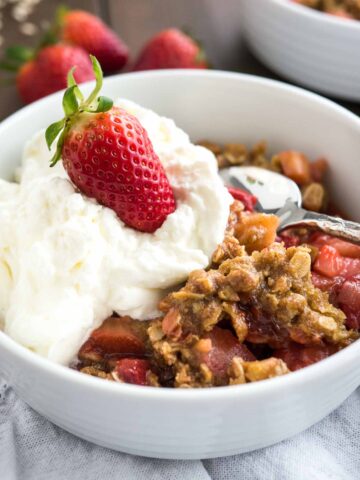 Strawberry rhubarb crisp in a white bowl topped with cream and a strawberry with a spoon in it.