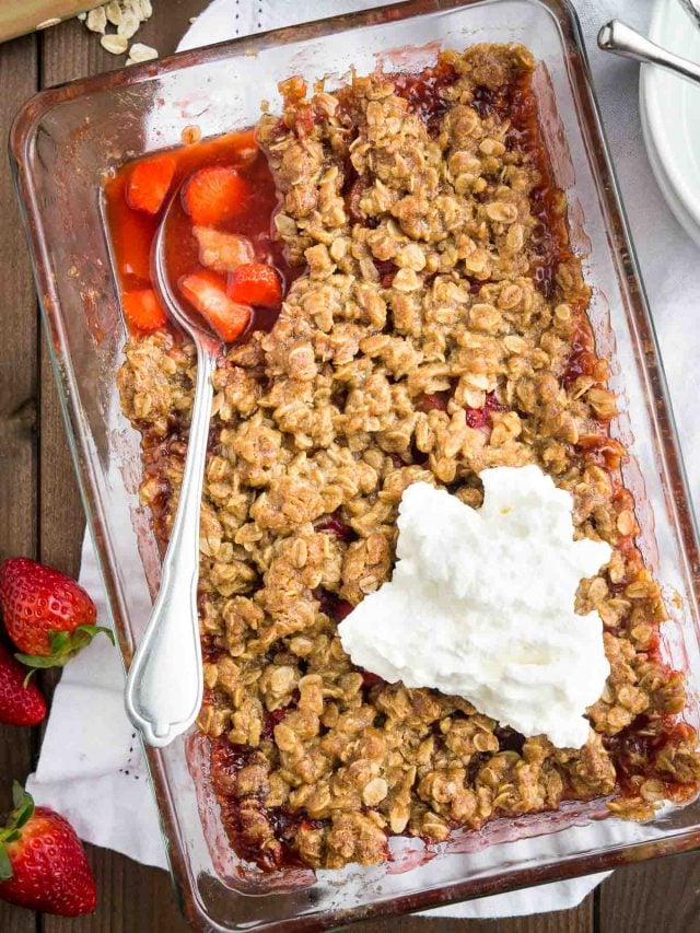 top-down shot of strawberry rhubarb crisp in a glass baking dish on a wooden table. It is topped with whipped cream and a spoon is digging into the strawberry rhubarb bottom layer