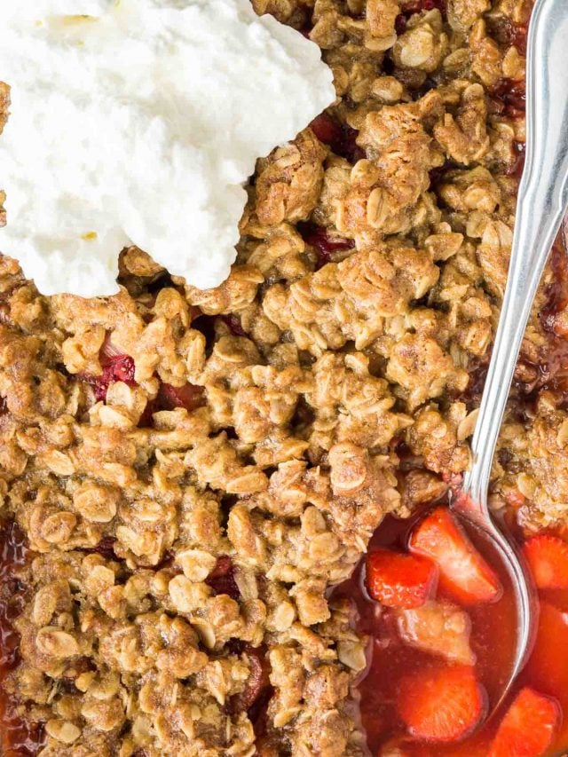 close up of strawberry rhubarb crisp topped with cream, with a spoon digging into the rhubarb-strawberry bottom layer