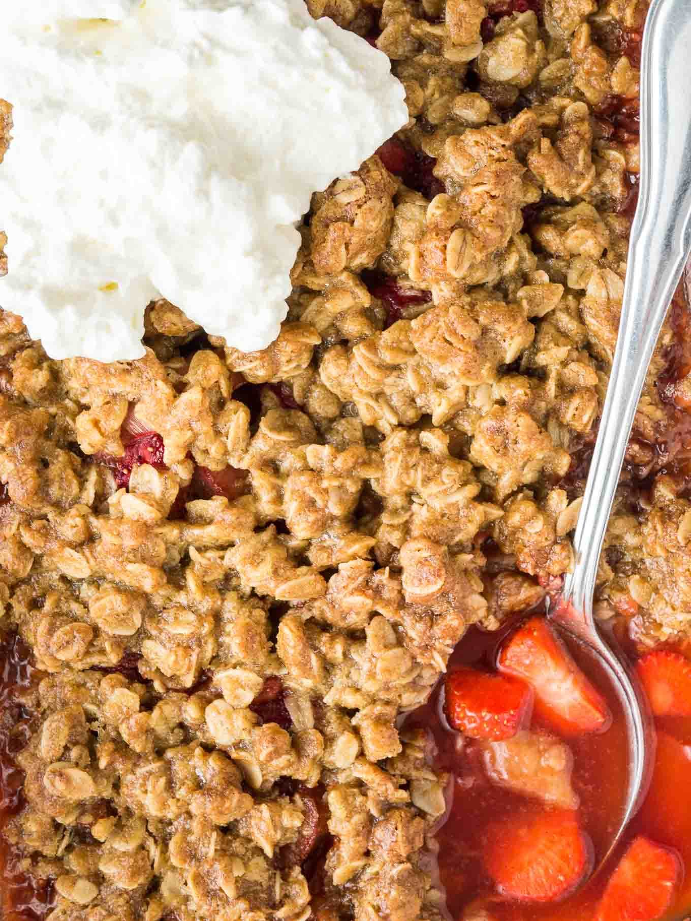 Strawberry rhubarb crisp topped with cream, with a spoon digging into the rhubarb-strawberry bottom layer.