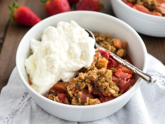 strawberry rhubarb crisp in a white bowl with a spoon in it on a wooden table.  it is topped with whipped cream and there are fresh strawberries in the background.