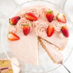Top-down shot of a strawberry cake topped with strawberries on a glass serving dish with a slice being lifted out with a cake server.