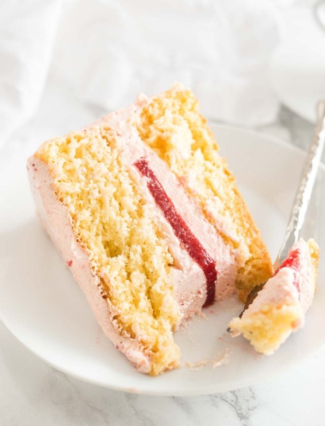 A slice of strawberry cake on a white plate. A fork is taking out a piece of the slice.