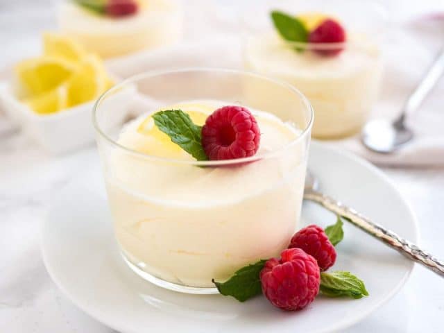 A glass of lemon mousse, garnished with mint, raspberries and lemon slices on a white plate with a spoon. There is a white bowl with lemon slices and more glasses of mousse in the background.