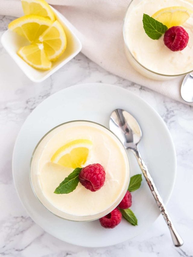 Top-down view of lemon mousse in a glass on a white plate with a spoon garnished with mint, lemon slices and raspberry, next to a small square bowl of lemon slices and another glass of mousse.