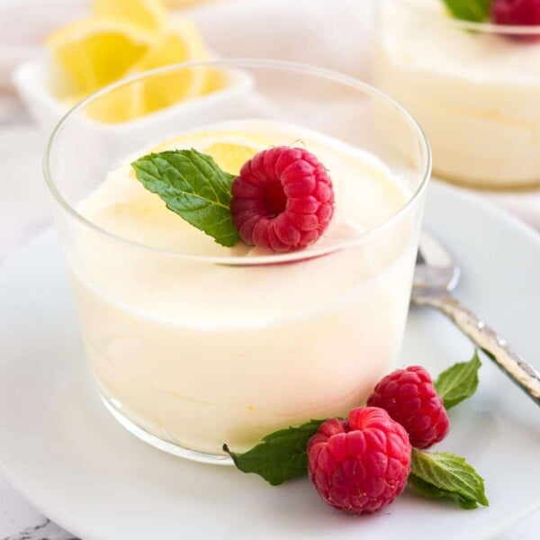 Close-up of a glass of lemon mousse, garnished with mint, raspberries and lemon slices on a white plate with a spoon. There is a white bowl with lemon slices and more glasses of mousse in the background.