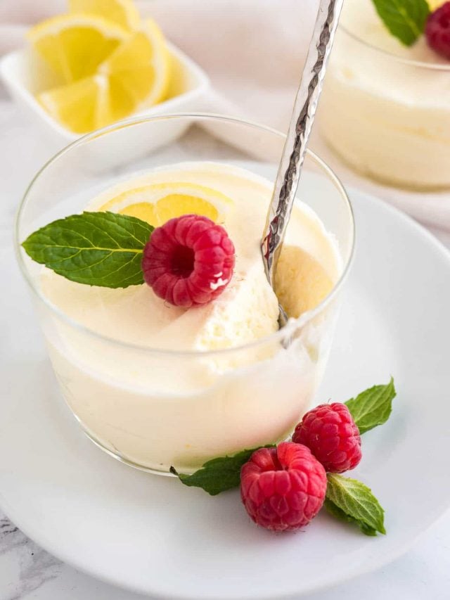 A glass of lemon mousse, garnished with mint, raspberries and lemon slices on a white plate with a spoon. There is a white bowl with lemon slices and more glasses of mousse in the background.