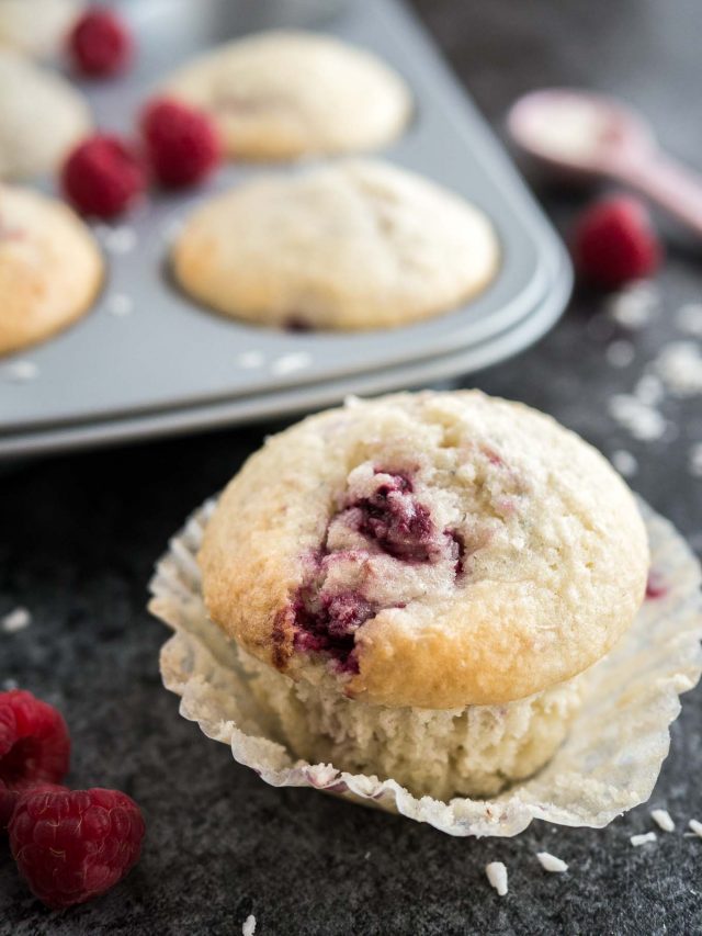 A coconut raspberry muffin with its liner opened sitting on a dark surface. There are raspberries, a pink measuring spoon with coconut flakes and a grey muffin pan with more muffins next to it.