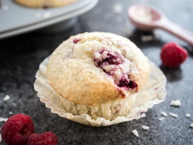 Close-up of a coconut raspberry muffin with its liner opened sitting on a dark surface. There are raspberries, a pink measuring spoon with coconut flakes and a grey muffin pan with more muffins next to it.