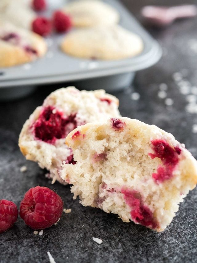 Close-up of a coconut raspberry muffin broken into two pieces garnished with raspberries. There\'s a grey muffin pan with more muffins in the background.