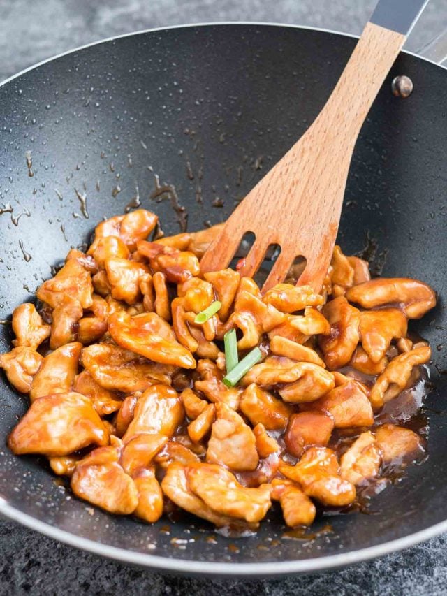 A Wok with general tso\'s chicken with a wooden spatula.