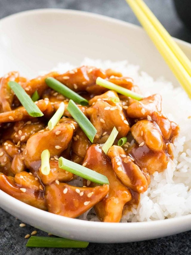 A grey bowl with rice and general tso\'s chicken, garnished with chives, with chopsticks.