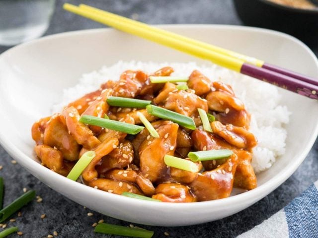 A grey bowl with rice and general tso\'s chicken, garnished with chives, with chopsticks on a dark surface with chives and sesame seeds.