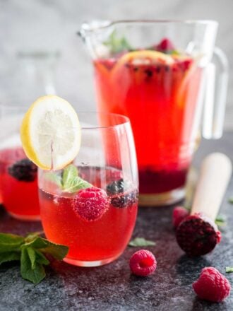 Two glasses of raspberry lemonade with raspberries and mint in them and a slice of lemon on the rim. There's a jug with the rest of the lemonade in the background and a muddler next to it.